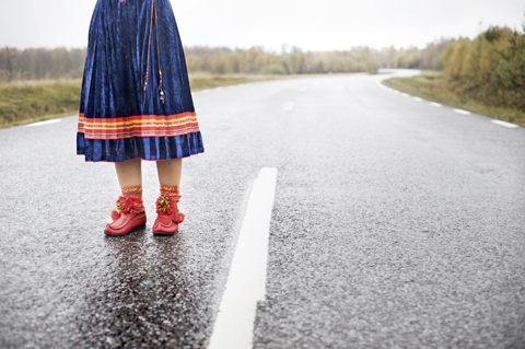 samisk flicka står på asfalterad väg. Hon bär röda näbbskor och blå samisk dräkt.