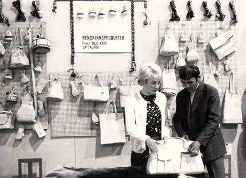 En gammal bild från Kero som visar en vägg med diverse handväskor och väskor. två personer inspekterar en väska.